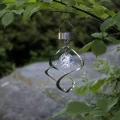 Solarna dekoracja wisząca ze stalową spiralą i ozdobną kulą świetlną, zawieszona na gałęzi wśród zielonych liści ogrodu.