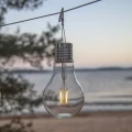 Solarna dekoracja LED w kształcie żarówki zawieszona na linkach nad wodą. Przezroczysta obudowa z widocznym filamentem emituje ciepłe światło, tworząc nastrojową ozdobę do ogrodu, na taras lub balkon.