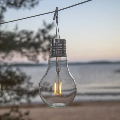 Solarna dekoracja LED w kształcie żarówki zawieszona na linkach nad wodą. Przezroczysta obudowa z widocznym filamentem emituje ciepłe światło, tworząc nastrojową ozdobę do ogrodu, na taras lub balkon.
