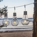 Solarne lampki LED w kształcie klasycznych żarówek zawieszone na lince. Przezroczyste obudowy z widocznymi filamentami emitują ciepłe światło, tworząc nastrojową dekorację ogrodu, tarasu lub balkonu.
