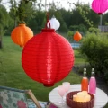 Czerwony lampion solarny LED zawieszony nad ogrodowym stolikiem wśród kolorowych lampionów. Dekoracja tworzy przytulną atmosferę podczas letnich spotkań na tarasie i w ogrodzie.