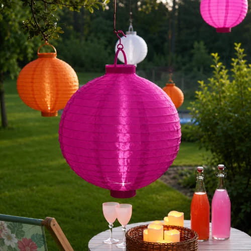 Wiszący różowy lampion solarny LED w papierowym kloszu zawieszony na gałęzi drzewa. Dekoracja ogrodowa tworząca przyjemny nastrój po zmroku.