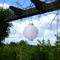 Biały lampion solarny LED zawieszony pod pergolą na tle zielonego ogrodu i błękitnego nieba. Dekoracja doskonale sprawdza się na tarasie, balkonie lub w altanie.