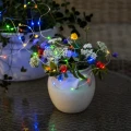 Kolorowy łańcuch solarny Dew Drop LED wykorzystany do dekoracji kompozycji kwiatowej w ceramicznym wazonie. Mikro diody w barwach czerwonej, zielonej, niebieskiej i żółtej tworzą nastrojowe oświetlenie stołu lub tarasu.