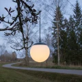Świecąca lampa solarna w formie mlecznej kuli zawieszona na łańcuszku. Dekoracyjne oświetlenie ogrodowe prezentowane na tle drzew i alejki o zmierzchu.