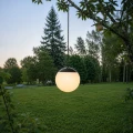 Kulista lampa solarna zawieszona na gałęzi drzewa nad trawnikiem. Mleczny klosz emituje ciepłe światło, a w tle widoczny jest ogród z wysokimi drzewami o zmierzchu.