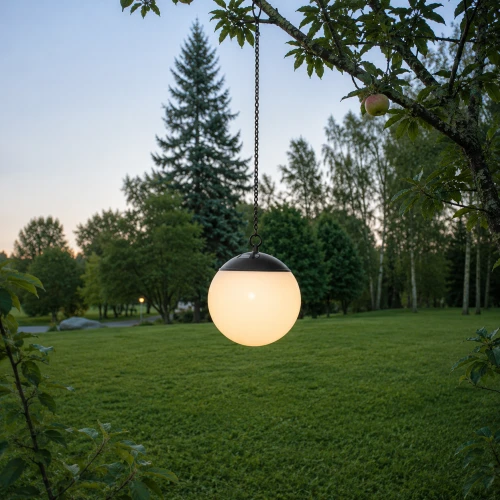 Kulista lampa solarna zawieszona na gałęzi drzewa nad trawnikiem. Mleczny klosz emituje ciepłe światło, a w tle widoczny jest ogród z wysokimi drzewami o zmierzchu.
