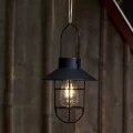 Czarna lampa solarna z dekoracyjnym daszkiem i osłoną żarówki emituje ciepłe światło na tle drewnianej ściany, tworząc przytulny nastrój po zmroku.