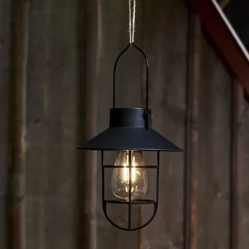 Czarna lampa solarna z dekoracyjnym daszkiem i osłoną żarówki emituje ciepłe światło na tle drewnianej ściany, tworząc przytulny nastrój po zmroku.