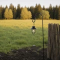 Wisząca lampa solarna umieszczona na smukłym stojaku ogrodowym stanowi subtelny punkt świetlny na tle zielonej łąki i drewnianego płotu.