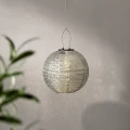 Wiszący lampion solarny LED w kształcie kuli z ażurową osłoną zawieszony na cienkiej lince. Delikatnie rozproszone światło podkreśla perforowany wzór i tworzy subtelne refleksy na ścianie.