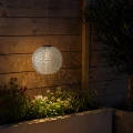 Wiszący lampion solarny LED w kształcie kuli oświetla rabatę przy drewnianej ścianie. Ażurowa osłona tworzy dekoracyjne refleksy świetlne na deskach i wśród roślin po zmroku.