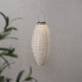 Ażurowy lampion solarny LED o podłużnym kształcie zawieszony na cienkiej lince. Delikatnie rozprasza światło, tworząc subtelne refleksy na jasnej ścianie.