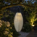 Podłużny lampion solarny LED zawieszony na gałęzi drzewa w ogrodzie. Ciepłe światło podkreśla rośliny i tworzy dekoracyjne refleksy po zmroku.