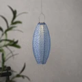 Podłużny lampion solarny LED zawieszony na cienkiej lince. Ażurowa powierzchnia delikatnie rozprasza światło i tworzy subtelne refleksy na jasnej ścianie.