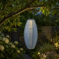 Podłużny lampion solarny LED zawieszony na gałęzi drzewa w ogrodzie. Ciepłe światło podkreśla rośliny i tworzy przytulny nastrój po zmroku.