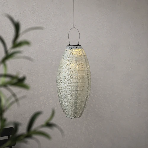 Podłużny lampion solarny LED zawieszony na cienkiej lince. Ażurowa powierzchnia delikatnie rozprasza światło, tworząc subtelny wzór na jasnej ścianie.