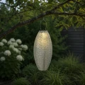 Lampion solarny LED zawieszony na gałęzi drzewa w ogrodzie. Ciepłe światło podkreśla roślinność i tworzy przyjemną atmosferę po zmroku.