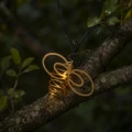 Zbliżenie na metalową dekorację LED w kształcie pszczoły zamocowaną na przewodzie łańcucha solarnego, ukazujące spiralny korpus i ciepłe światło diody.