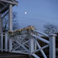 Girlanda z zielonych gałązek i ciepłobiałymi lampkami LED ułożona wzdłuż białej balustrady. Zwisające pędy podkreślają linię poręczy na tle wieczornego nieba i zimowego krajobrazu.