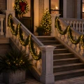 Zielona girlanda świąteczna z ciepłobiałymi lampkami LED ozdabiająca balustrady schodów prowadzących do wejścia domu. Tworzy elegancką, świąteczną aranżację z wiankiem i choinką.
