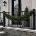Girlanda z gęstego zielonego igliwia ozdabia czarną metalową balustradę przy wejściu do domu. W tle widoczne są przeszklone drzwi, latarnia ścienna i choinka w donicy.