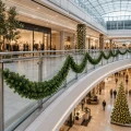 Zbliżenie na zieloną girlandę świąteczną zawieszoną na szklanej balustradzie galerii handlowej. W tle znajdują się podświetlone kolumny, sklepy oraz choinka na niższym poziomie.