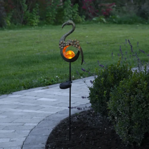 Solarna dekoracja ogrodowa w kształcie flaminga z podświetlaną bursztynową kulą umieszczona przy ścieżce. W tle widoczny jest trawnik oraz przycięte krzewy w ogrodowej aranżacji.