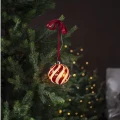 Bombka LED z czerwonym spiralnym wzorem zawieszona na gałązce choinki. Rozmyte punkty światła w tle podkreślają świąteczną aranżację.