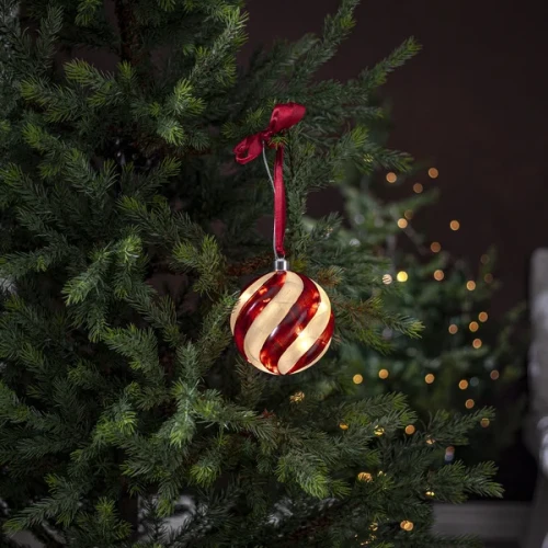 Szklana bombka LED z czerwonym spiralnym wzorem zawieszona na gałązce choinki na czerwonej satynowej wstążce. Ciemne tło podkreśla ciepłe światło dekoracji.