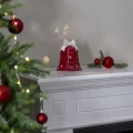 Na białej komodzie ustawiono czerwoną ceramiczną dekorację LED w kształcie dzwonka z białą kokardą. Obok leżą czerwone bombki i gałązka iglasta, a w tle widoczna jest choinka.
