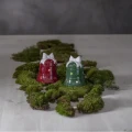 Dwa ceramiczne świeczniki w kształcie dzwonków w zielonym i czerwonym kolorze ustawiono obok siebie na jasnym blacie. Białe kokardy i wycięcia w kształcie gwiazdek podkreślają świąteczny charakter dekoracji, a w tle widoczny jest naturalny mech.