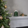 Zielony ceramiczny świecznik w kształcie dzwonka z białą kokardą ustawiony na białym kominku. W świątecznej aranżacji obok znajdują się gałązki i złote bombki, a z lewej strony widoczna jest udekorowana choinka.