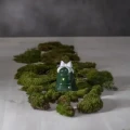 Na jasnym blacie, pośród naturalnego mchu, ustawiono zielony ceramiczny świecznik w kształcie dzwonka z białą kokardą. Wycięcia w formie gwiazdek subtelnie przepuszczają światło, tworząc spokojną świąteczną kompozycję.