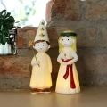 Przy ceglanej ścianie ustawiono dwie świecące figurki w białych strojach. Jedna trzyma złotą gwiazdkę, a druga świecę na czerwonej podstawce.