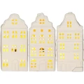 Białe ceramiczne domki dekoracyjne o trzech różnych kształtach, z licznymi okienkami rozświetlonymi ciepłym światłem.
