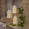 Trzy białe ceramiczne domki z podświetlanymi oknami ustawione na ceglanych stopniach. Aranżację uzupełniają zielone pnące gałązki.