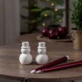Para białych świeczników bałwanków ustawiona na stole obok czerwonych świec, w tle świąteczna aranżacja z choinką i czerwonymi jabłkami.