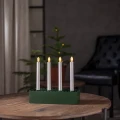 Zielony świecznik LED z czterema smukłymi świecami ustawiony na drewnianym stole, z małą choinką i ciemnym fotelem w tle.