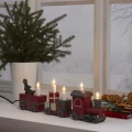 Świecznik dekoracyjny w formie świątecznego pociągu z pięcioma białymi świecami, ustawiony na parapecie obok zielonego drzewka i zimowego okna.