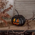Świecąca czarna dynia Halloween ustawiona na drewnianej półce w aranżacji z suchymi liśćmi, gałązkami i jasnym tłem.