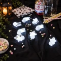 Świecące duszki na czarnym przewodzie ułożone na ciemnym materiale w halloweenowej aranżacji z kolorowymi piankami i dodatkami.