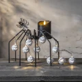 Łańcuch świetlny z białymi lampkami w formie dyń Halloween ułożony na metalowej dekoracji pająka, obok czarnej świecy na drewnianym blacie.
