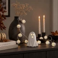 Łańcuch świetlny z białymi lampkami w formie dyń ułożony na ciemnej komodzie w halloweenowej aranżacji z dekoracjami duchów i świecami.