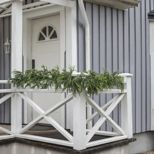 Zielona girlanda dekoracyjna ułożona na białej balustradzie przed wejściem do domu.
