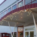 Trzy wiszące dekoracje świetlne w formie gwiazd rozmieszczone pod zadaszeniem wejścia, połączone falującą linią świetlną biegnącą wzdłuż krawędzi budynku.