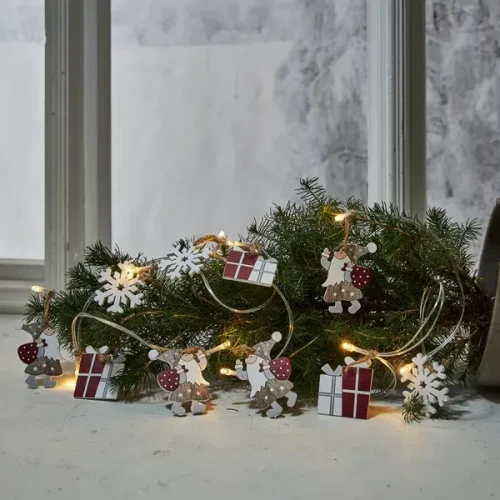 Łańcuch świąteczny LED z drewnianymi ozdobami przedstawiającymi Mikołaje z workami prezentów, płatki śniegu i paczki, ułożony wśród zielonych gałązek iglastych na parapecie.