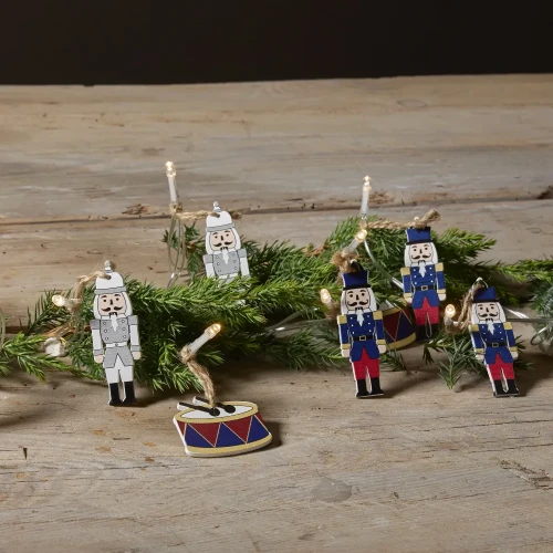 Świąteczny łańcuch LED z figurkami dziadków do orzechów w granatowych i szarych mundurach oraz ozdobnymi bębenkami, ułożony wśród zielonych gałązek na drewnianym blacie.