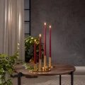 Elegancka kompozycja czterech smukłych świec LED w czerwonym kolorze, ustawionych na złotych podstawkach na drewnianym stoliku, z realistycznymi płomieniami świetlnymi i zielonymi roślinami w otoczeniu.