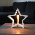 Dekoracyjna lampa stołowa w kształcie gwiazdy z białą ramą i żarówką LED E14 emitującą ciepłe światło na stoliku.